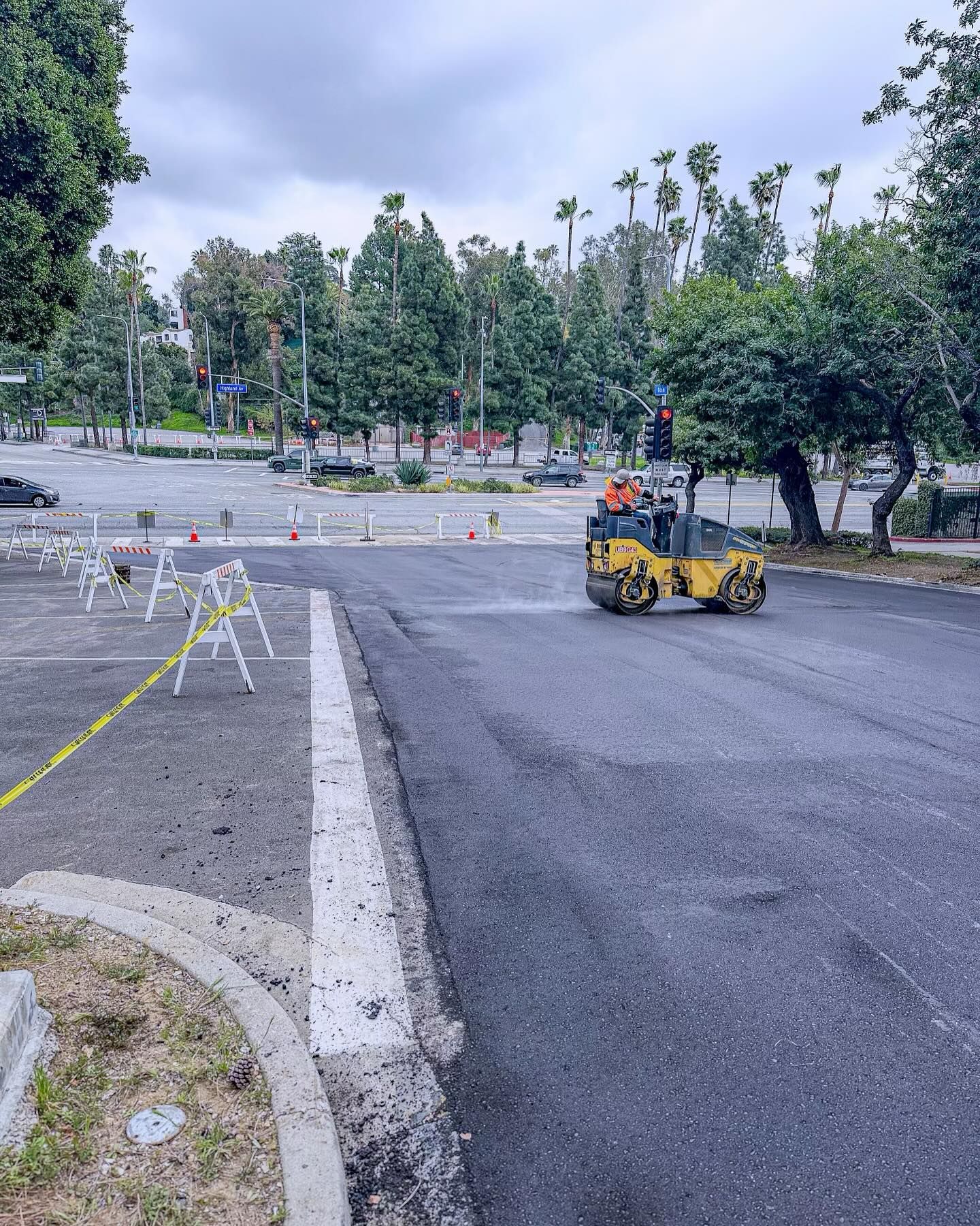 Freshly paved road with a small yellow road roller and traffic barriers in a tree-lined parking area