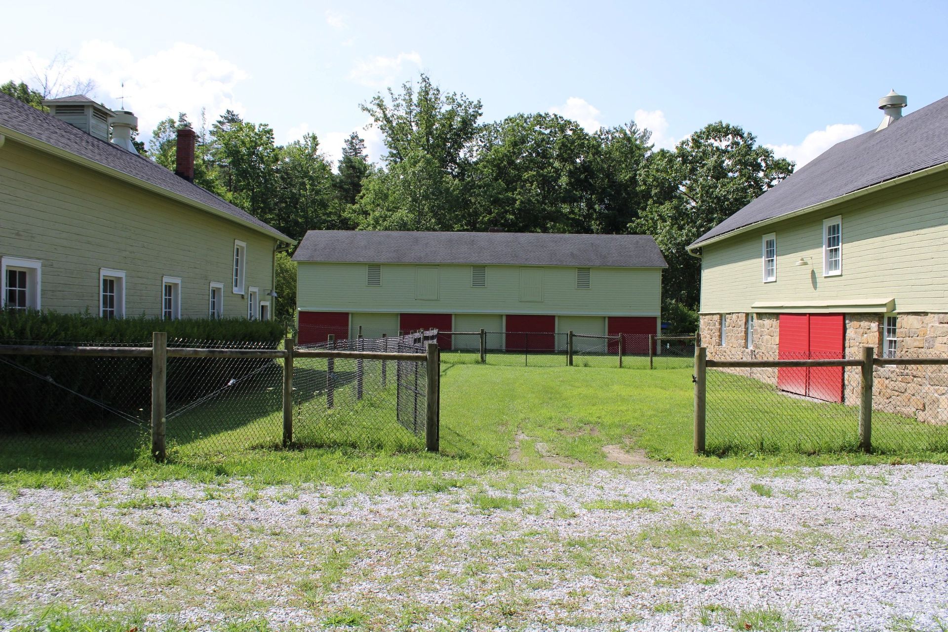 Green and red barns behind a chain link fence, gravel driveway, and a grassy yard.