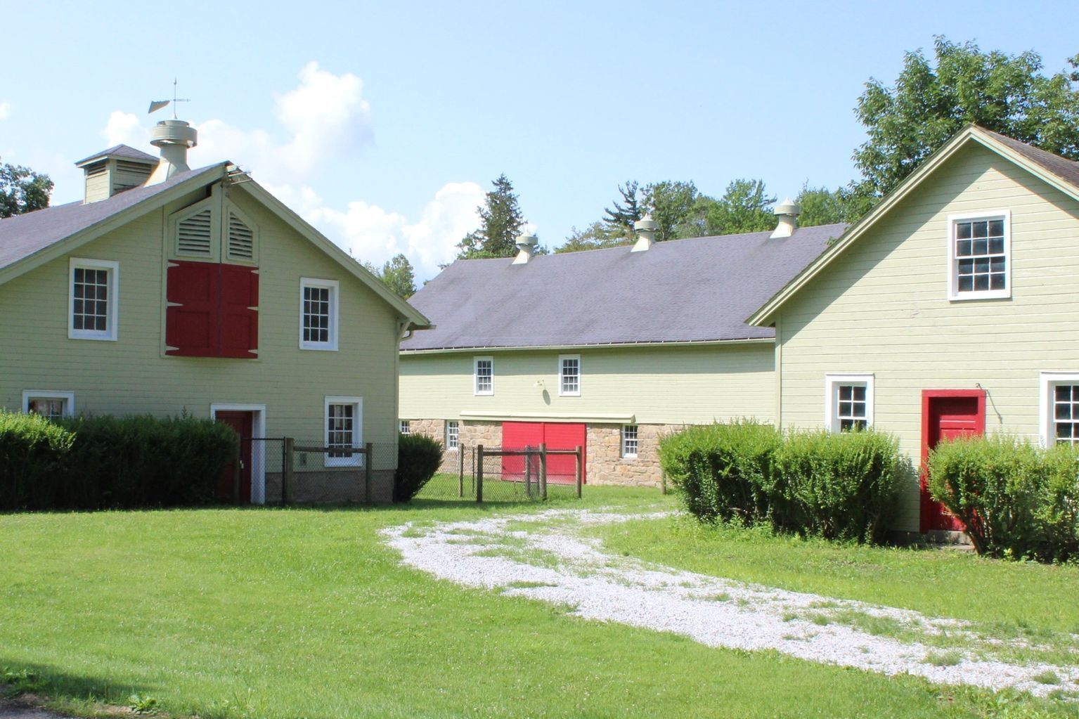 Green farm buildings with red doors and shutters on a sunny day. A gravel path leads to them.