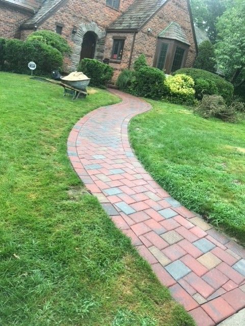 A brick walkway leading to a house with a wheelbarrow in the grass.