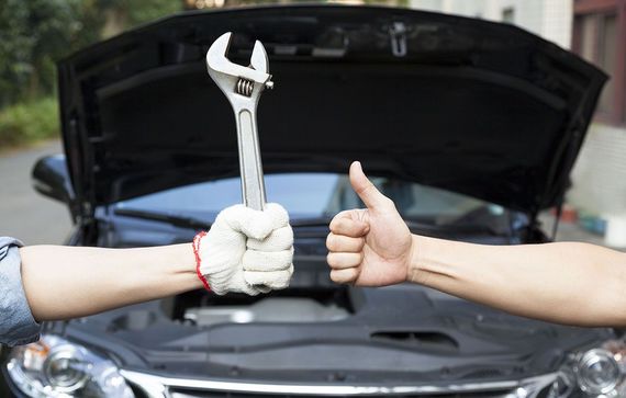 Two gloved hands under a car hood, one holding a wrench and the other giving a thumbs-up.