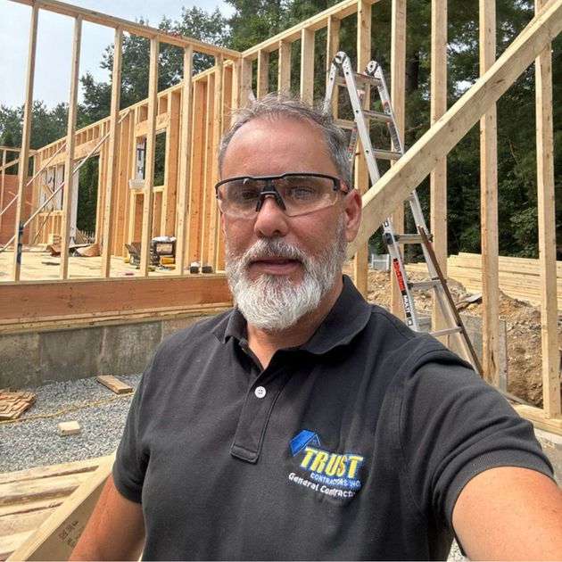 A construction worker wearing safety glasses and a logo polo shirt stands in front of a wooden building frame.