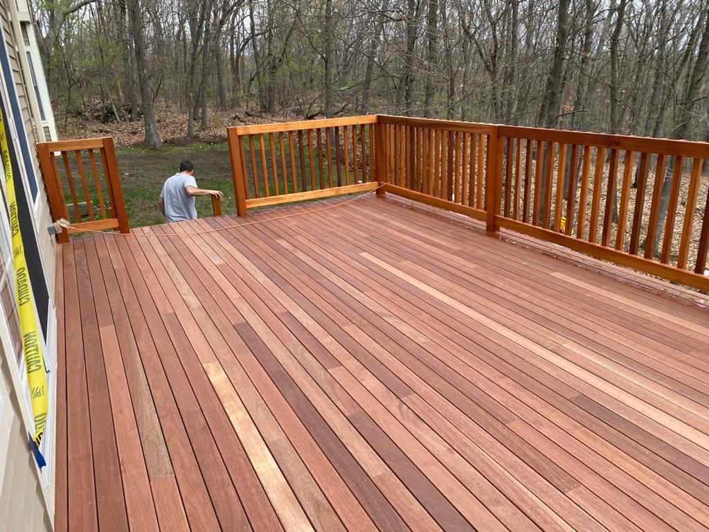 A person stands at the edge of a newly installed wooden deck with brown railings, overlooking a wooded yard.