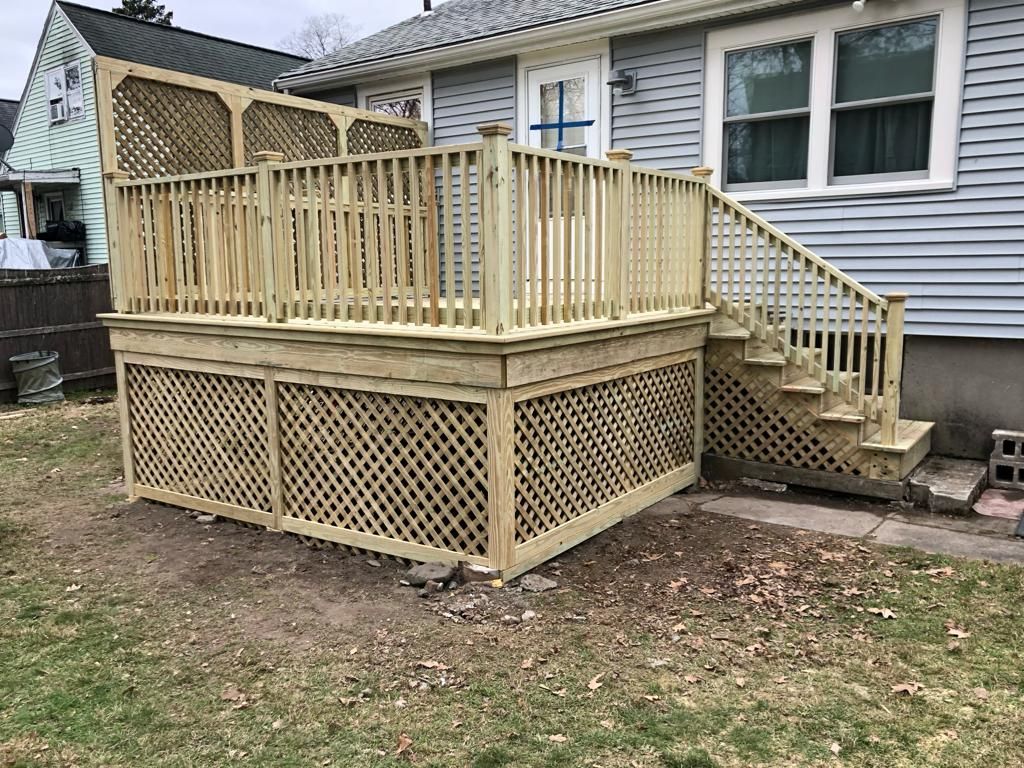 A newly built wooden deck with latticework skirting and railing attached to the back of a house with grey siding.
