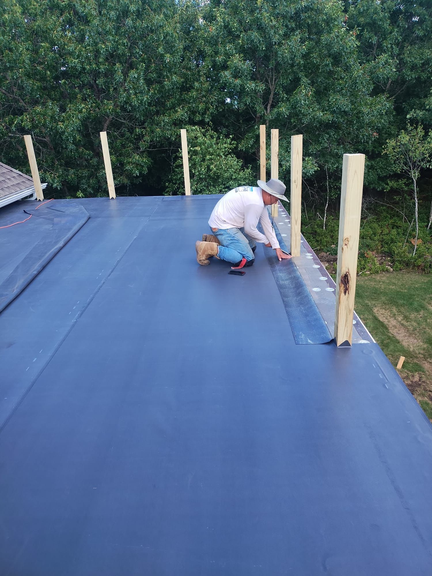 A person in a hat and white shirt installs dark roofing membrane on a flat roof with wooden support posts.