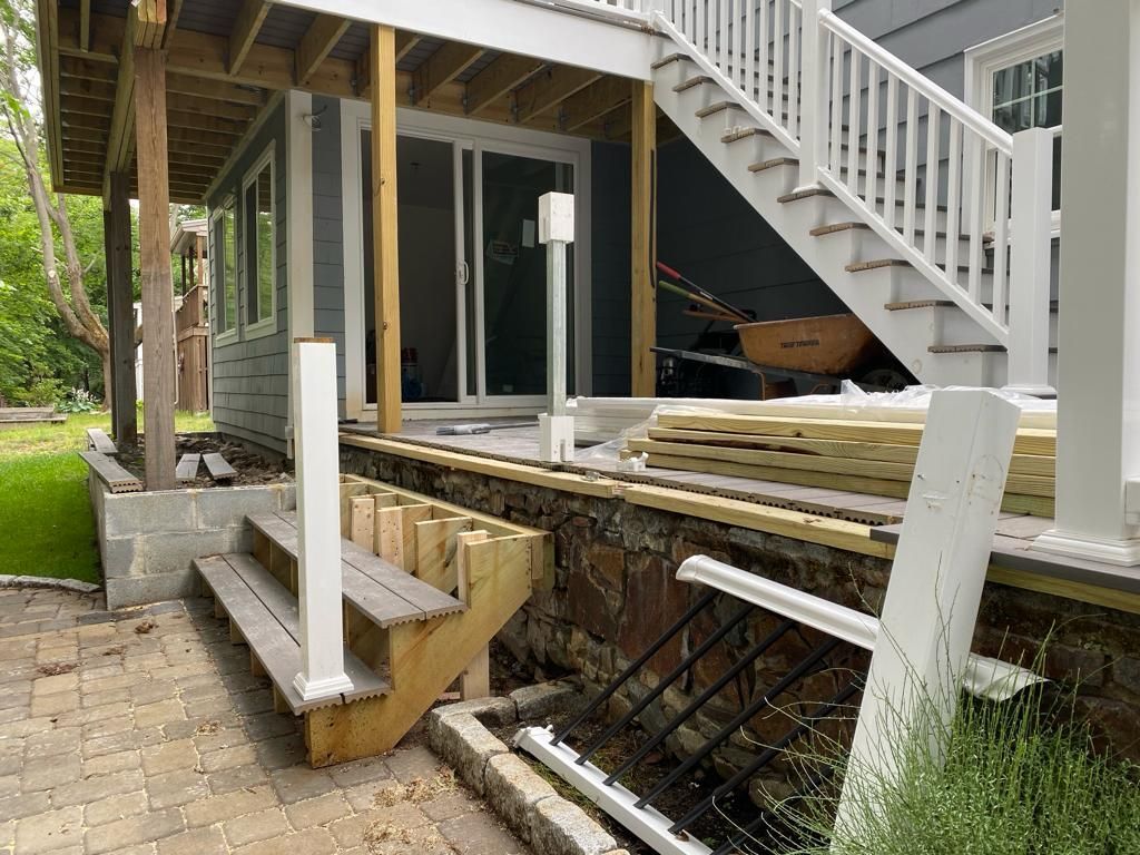 A partially constructed wooden deck and stairs under a white porch railing, set against a house with stone foundations.