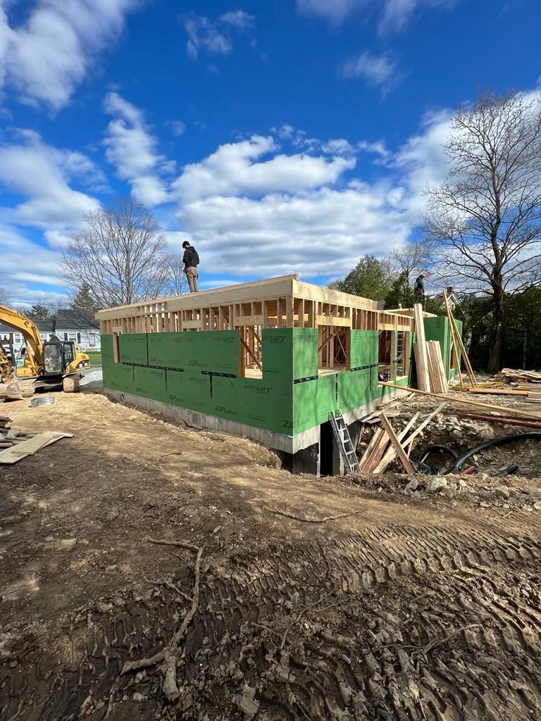 A construction site shows a partially built house with green wall sheathing, topped by wooden framing under a blue sky.