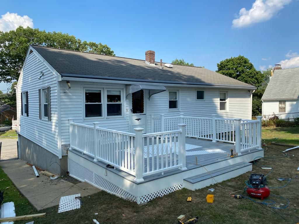 A white house with a newly constructed light-gray deck and white railing, surrounded by a yard under a clear blue sky.