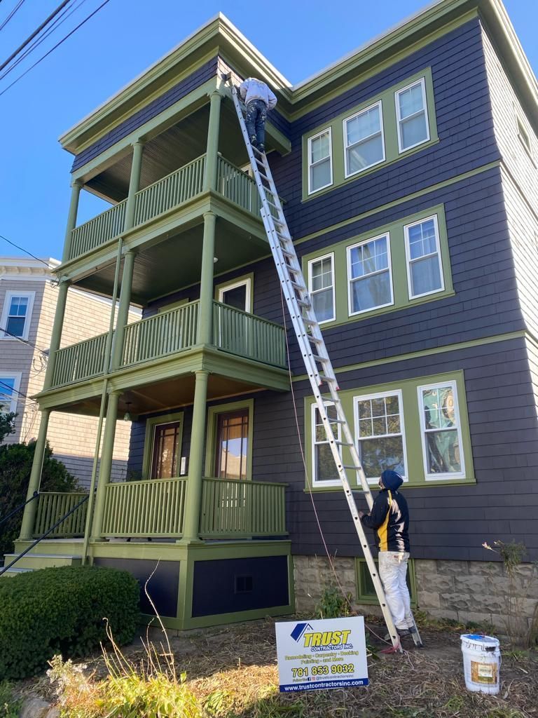 A worker on an extension ladder paints the trim of a three-story house with dark blue siding and green accents.