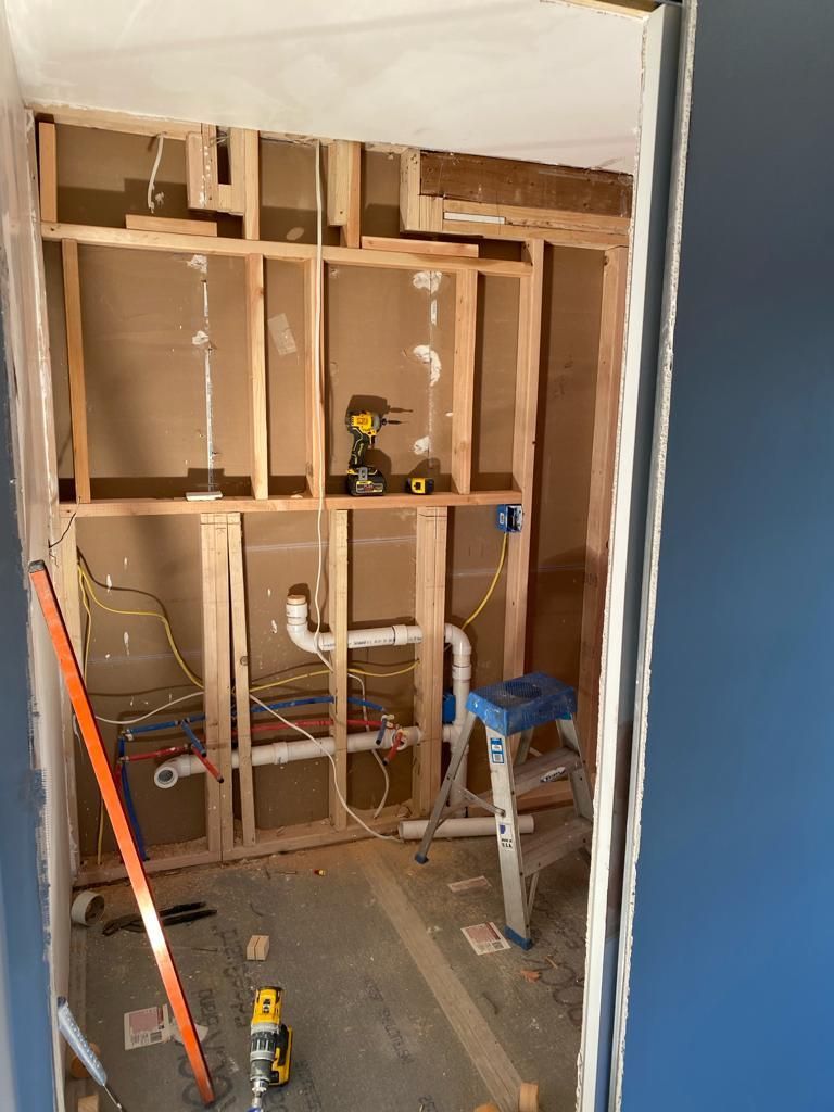 A wall cavity framed with wood studs, showing exposed plumbing, electrical wiring, and a stepladder.