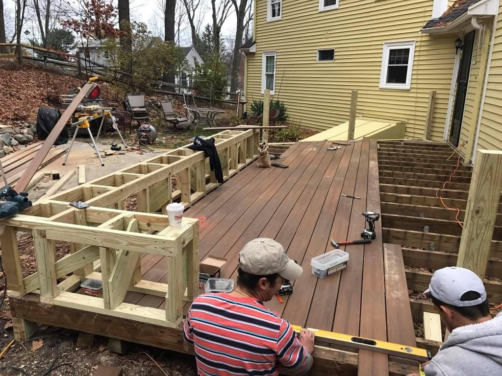 Two people wearing baseball caps construct a wooden deck attached to a yellow house, measuring boards for installation.
