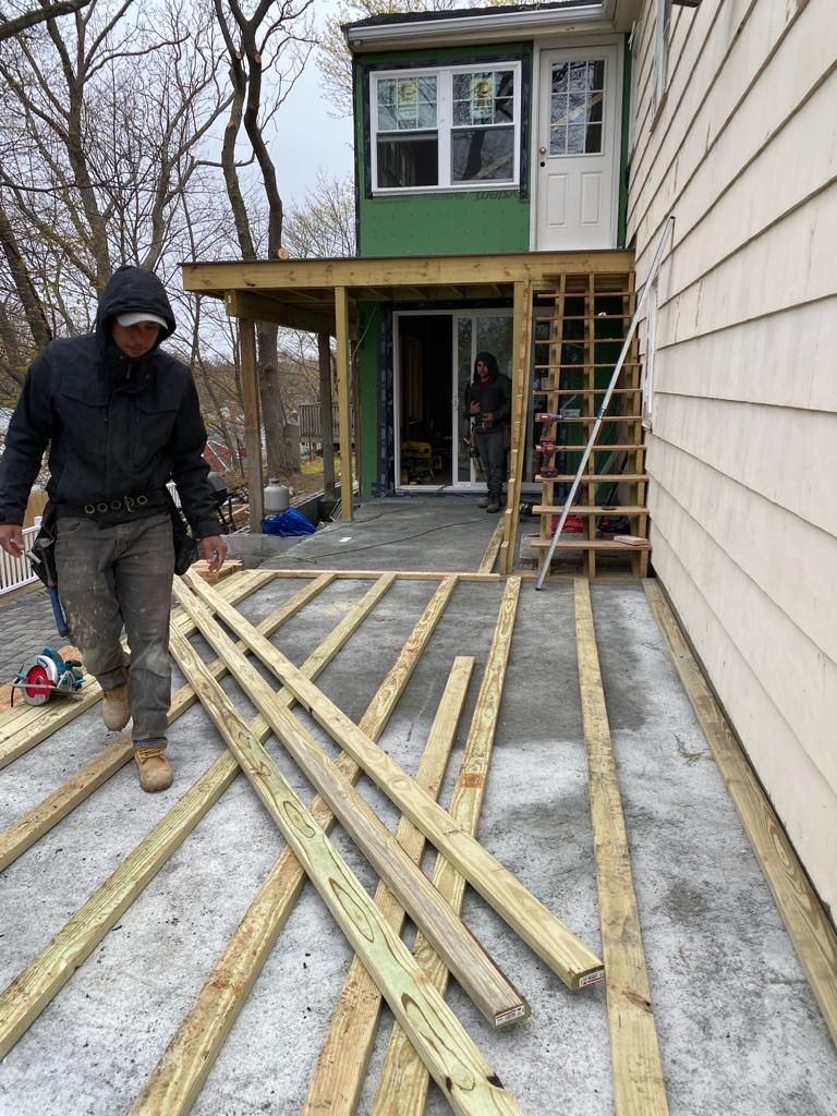 A construction worker in a black coat walks on a patio with laid-out wooden joists near a house with an outdoor staircase.
