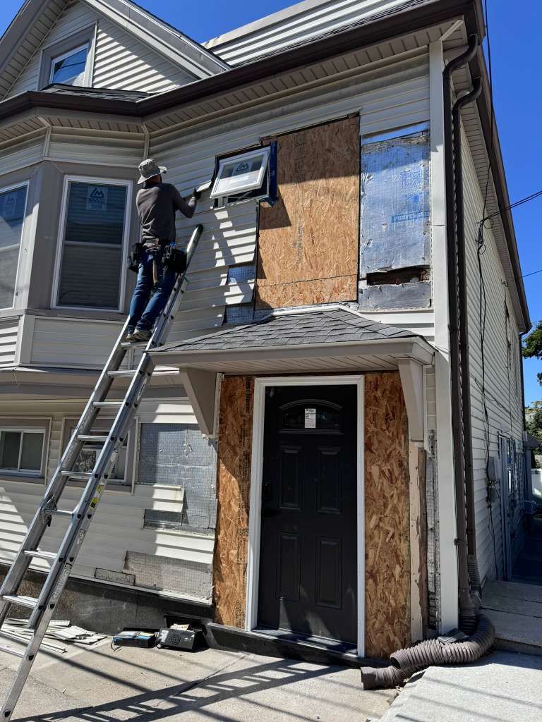 A person on an extension ladder works on the siding of a multi-story house with boarded-up windows.