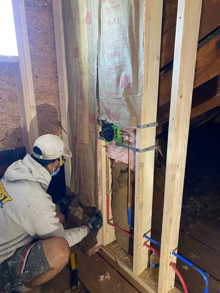 A construction worker wearing a cap and hoodie installs plumbing fixtures inside a wooden wall frame with insulation.
