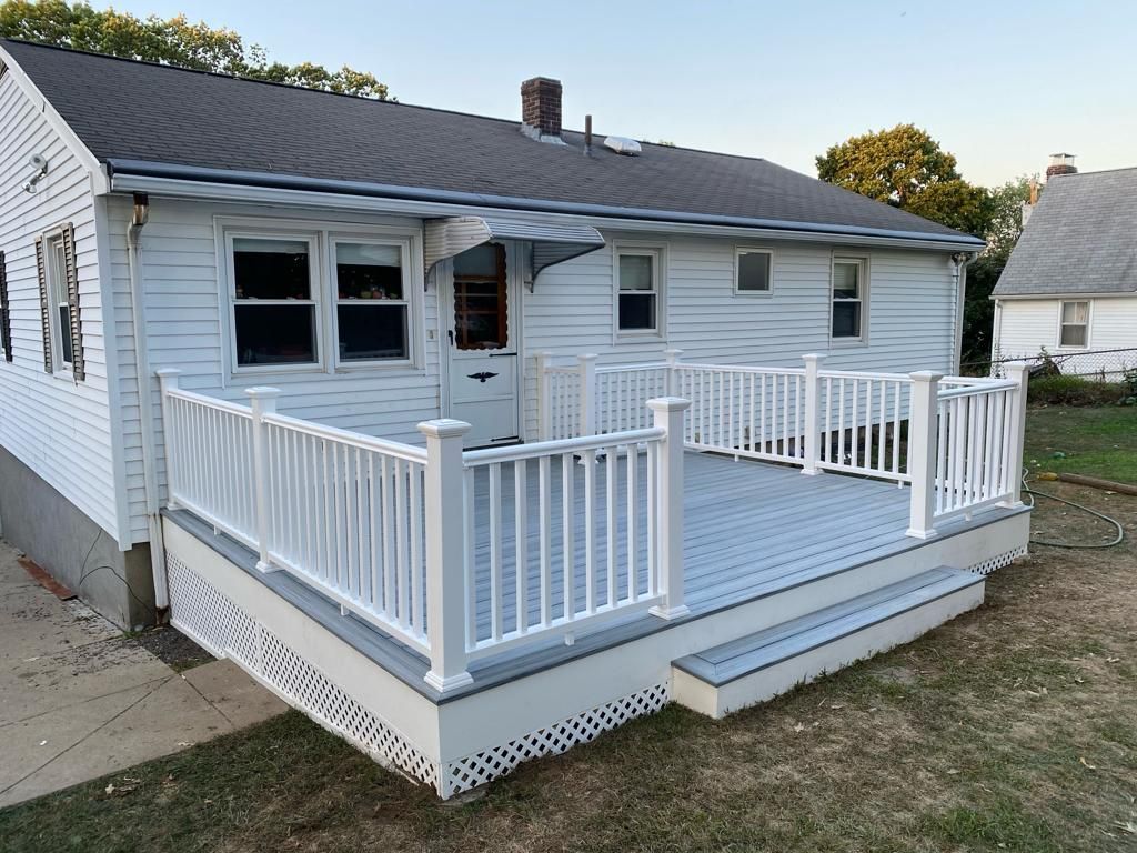 A white house with a gray deck, white railings, and lattice skirting, situated on a grass lawn during the daytime.