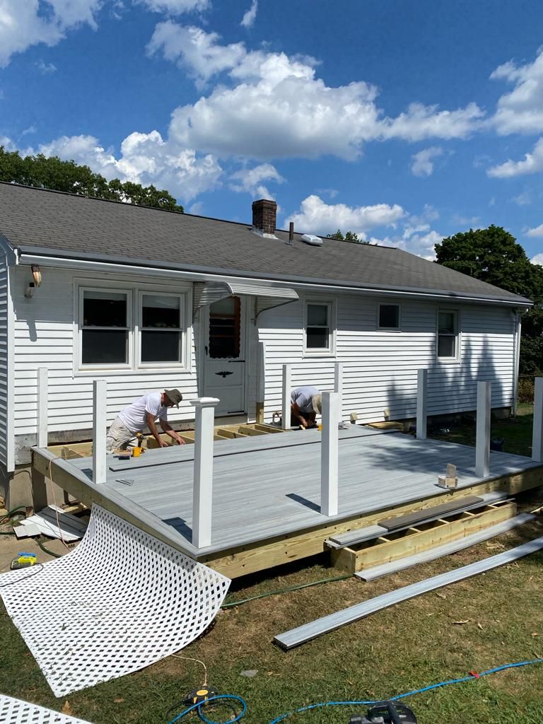 Two people work on a light gray deck being constructed on the back of a white house on a sunny day.