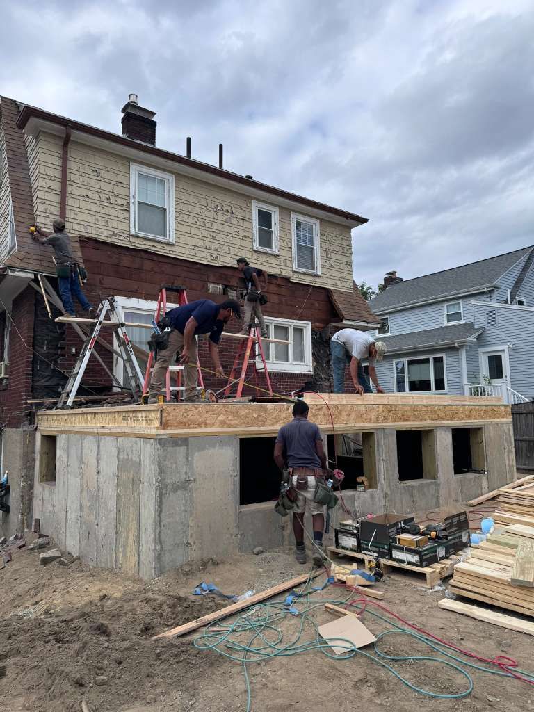 Construction crew builds a wooden deck onto the back of a brick house under a cloudy sky.