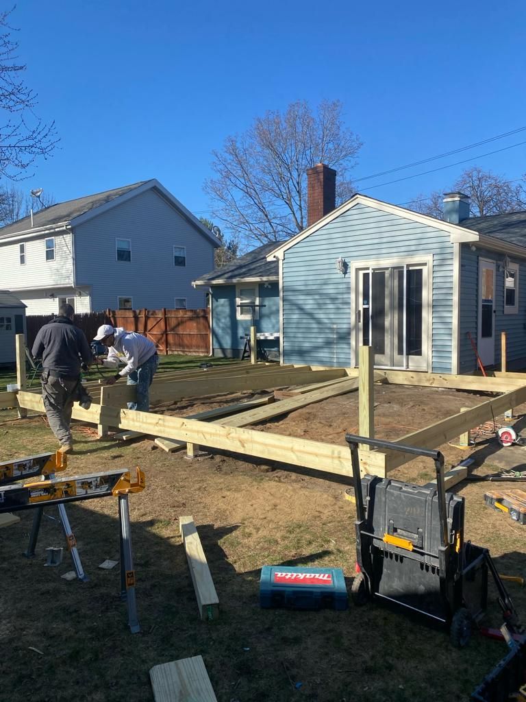 Two people work on a wooden deck frame in a backyard with a blue house in the background.