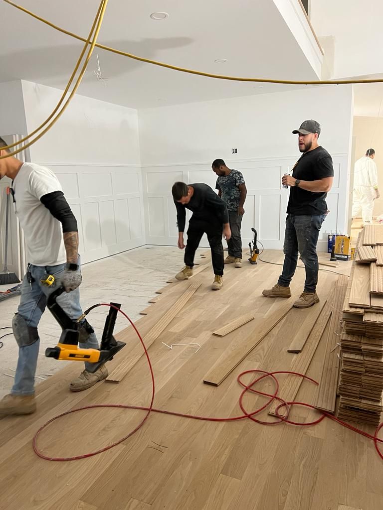 Four workers in a room with white walls and unfinished floors install light-colored hardwood flooring.
