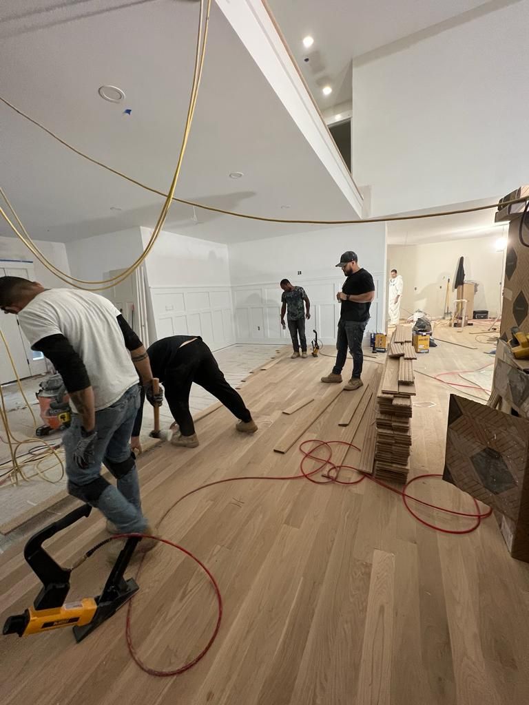 Construction workers install light-colored hardwood flooring in an unfinished room with white walls.