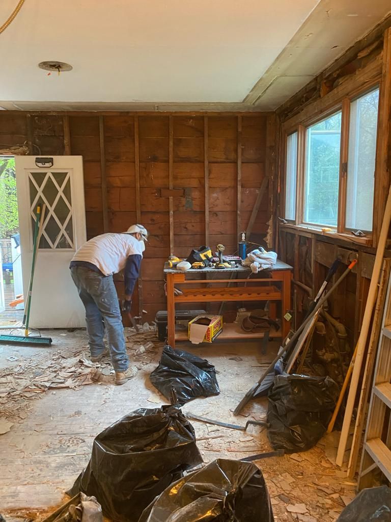 A worker in a room undergoing renovation, stripping wood wall paneling with piles of debris and tools on a workbench.