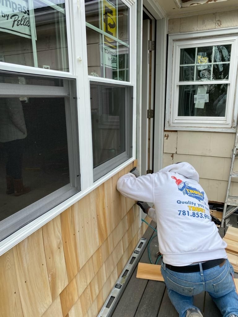 A person in a white hoodie installs cedar shingle siding below a window on an exterior wall using a level.