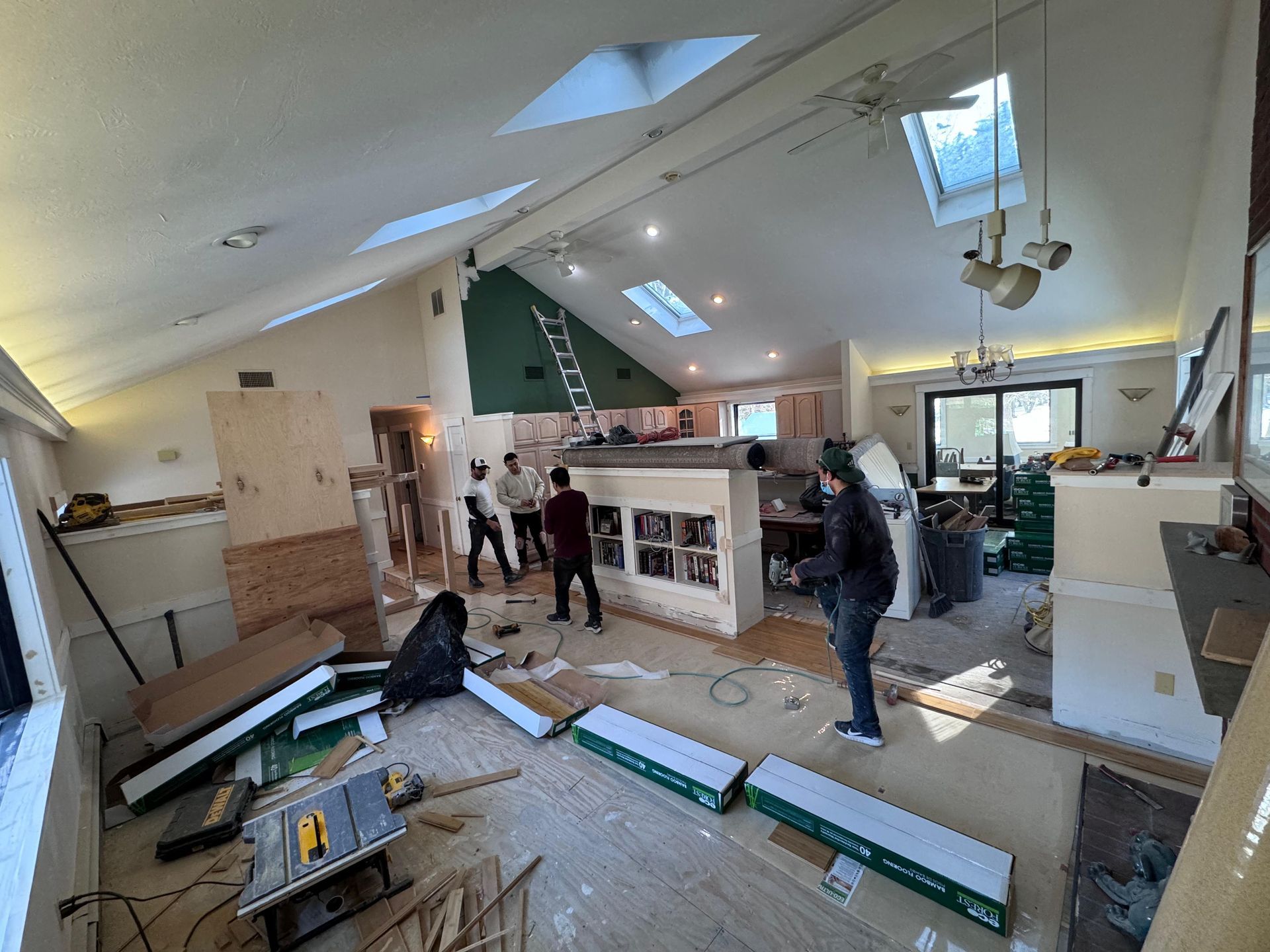 Workers renovate a room with vaulted ceilings and skylights, installing new flooring amidst tools and building materials.