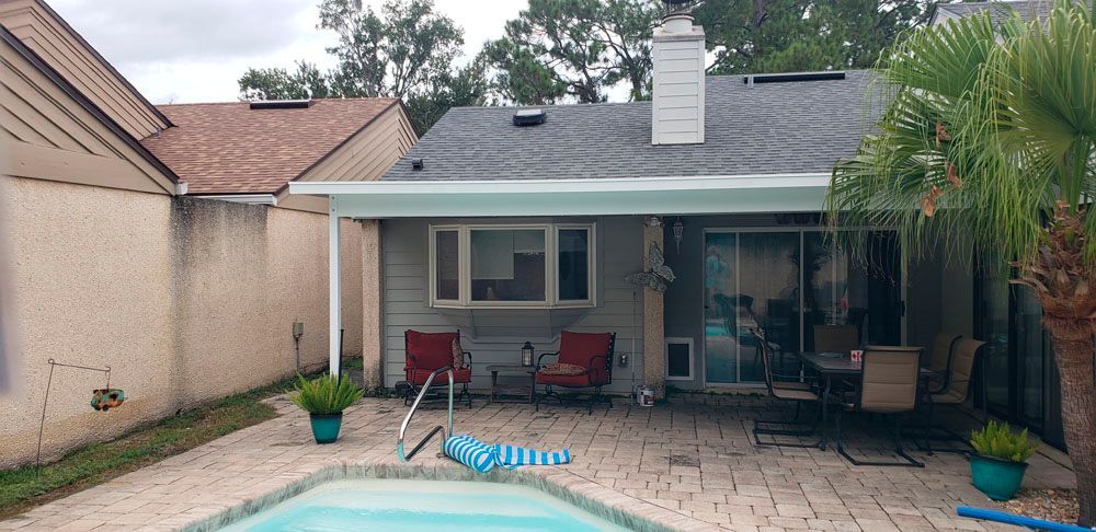 Backyard with a pool, patio, and house. There are chairs, a table, and a palm tree. Gray sky.