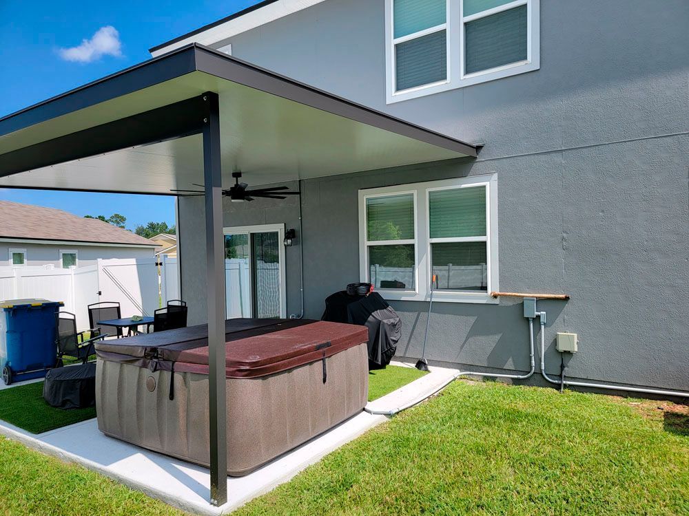 A patio with a hot tub under a canopy, near a house with gray walls and a window.