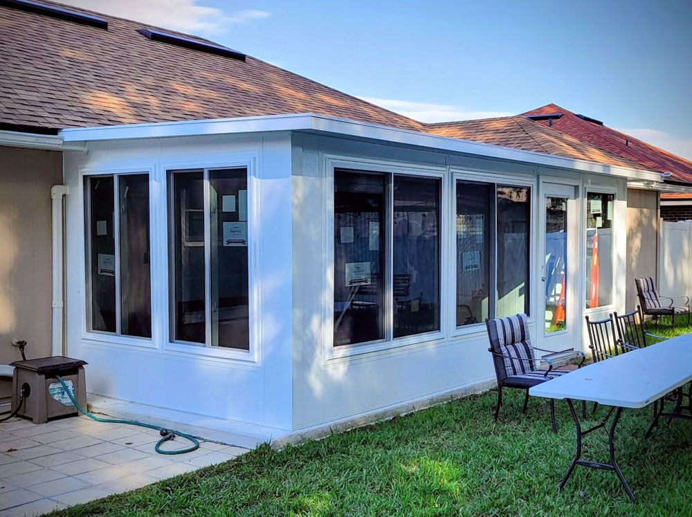 White sunroom addition attached to a house with windows and screens, on a patio and grassy area.