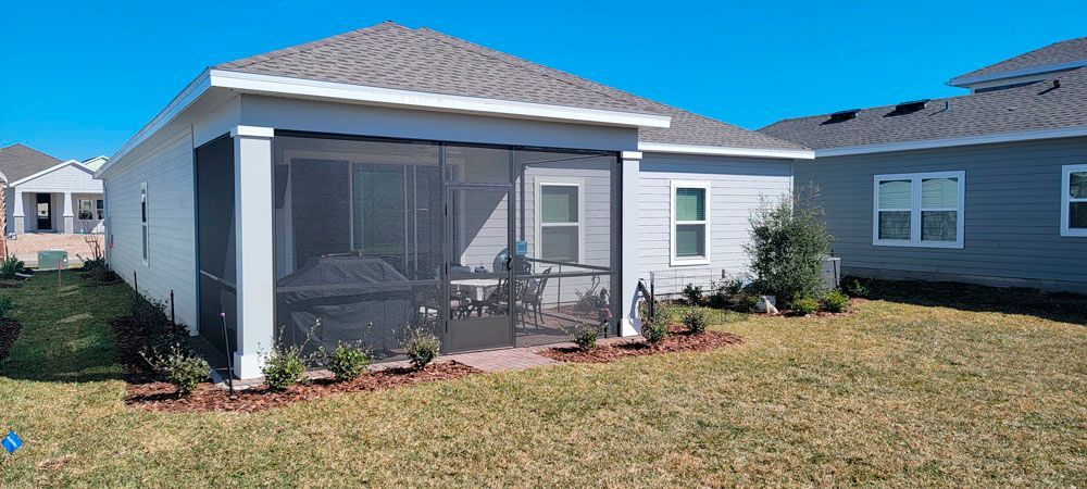 Backyard of a house with a screened-in porch and small landscaping in front of it. Blue sky.