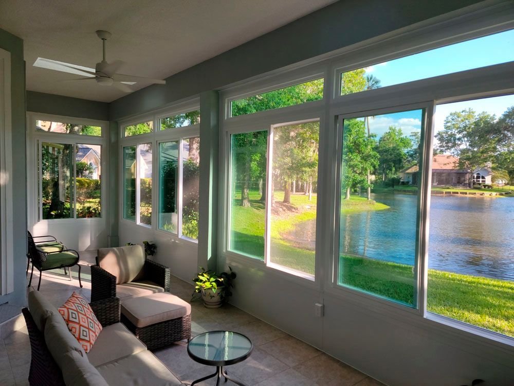 Sunroom with large windows overlooking a lake and green landscape; light-colored furniture.