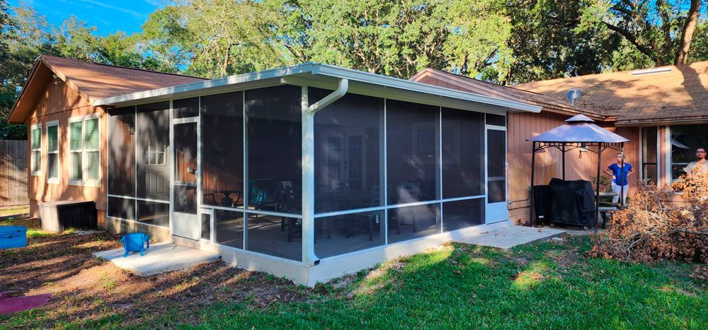 Screened porch attached to a brown house with a green lawn. Trees in the background.
