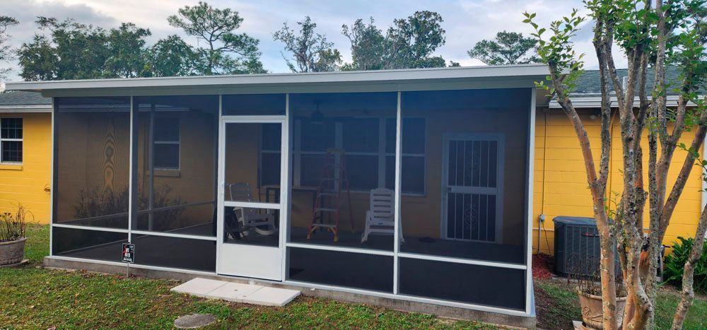 Screened-in porch on a yellow house with a white door. Green grass, trees in the background.