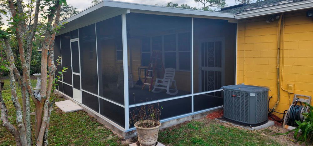 Screened-in porch attached to a yellow house with an air conditioning unit outside and chairs visible inside.