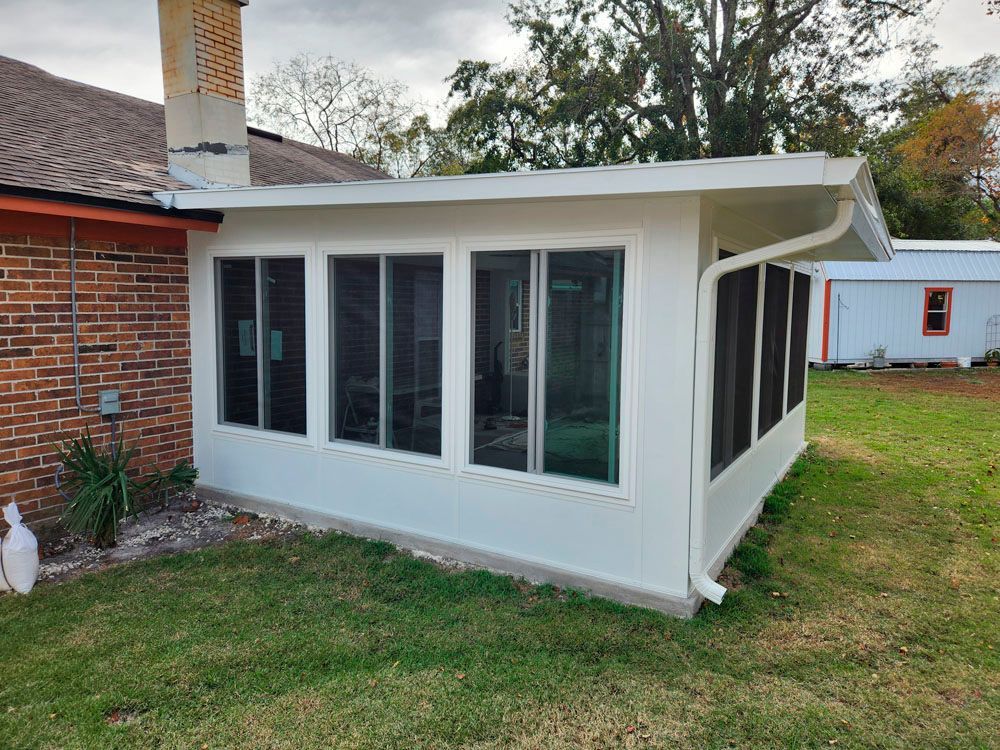 White screened-in porch attached to a brick house with chimney. Green grass surrounds it on an overcast day.
