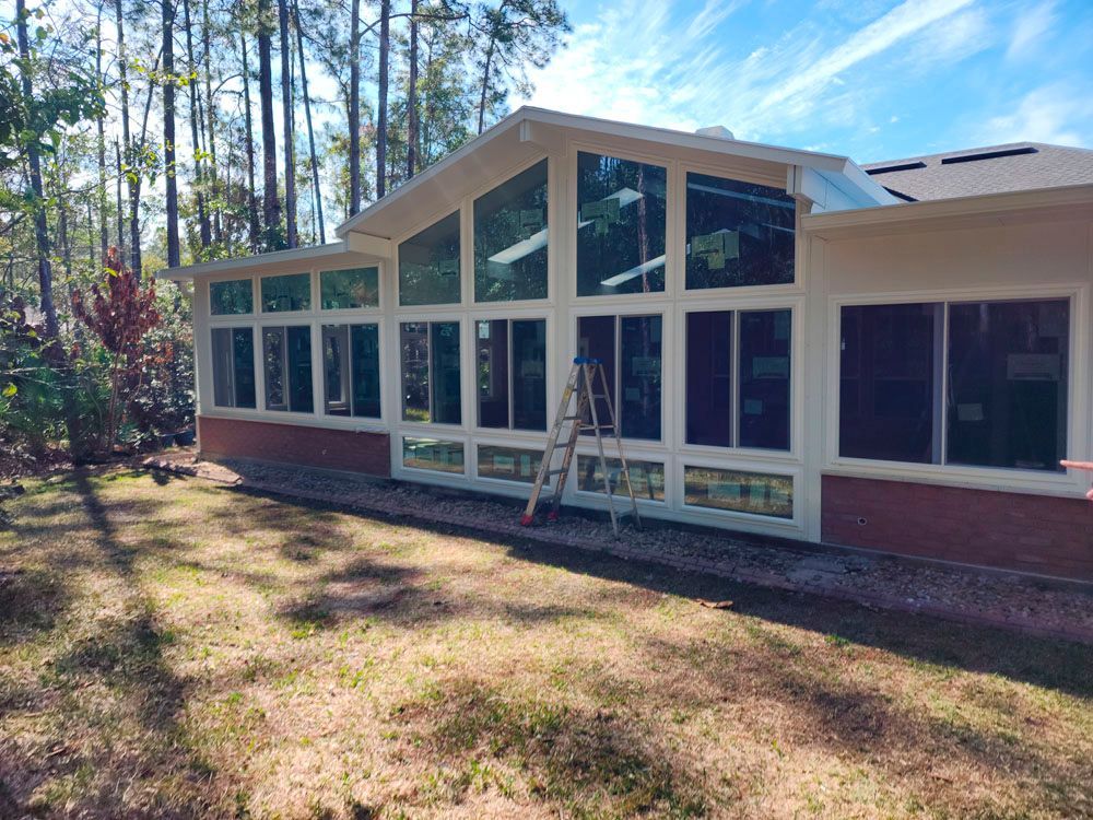 Sunroom with glass windows and a red brick base, surrounded by trees and grass on a sunny day.