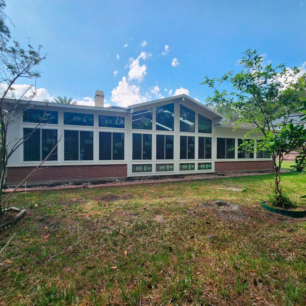 House with many windows, white walls, brick base, and a grassy yard under a blue sky with clouds.
