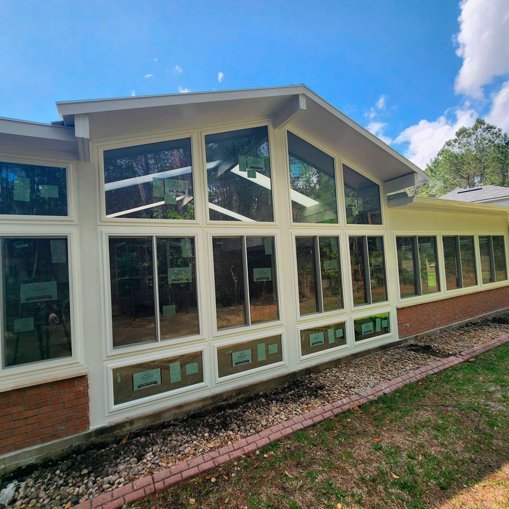 A sunroom with large windows, white frames, brick base, and a gabled roof against a blue sky.