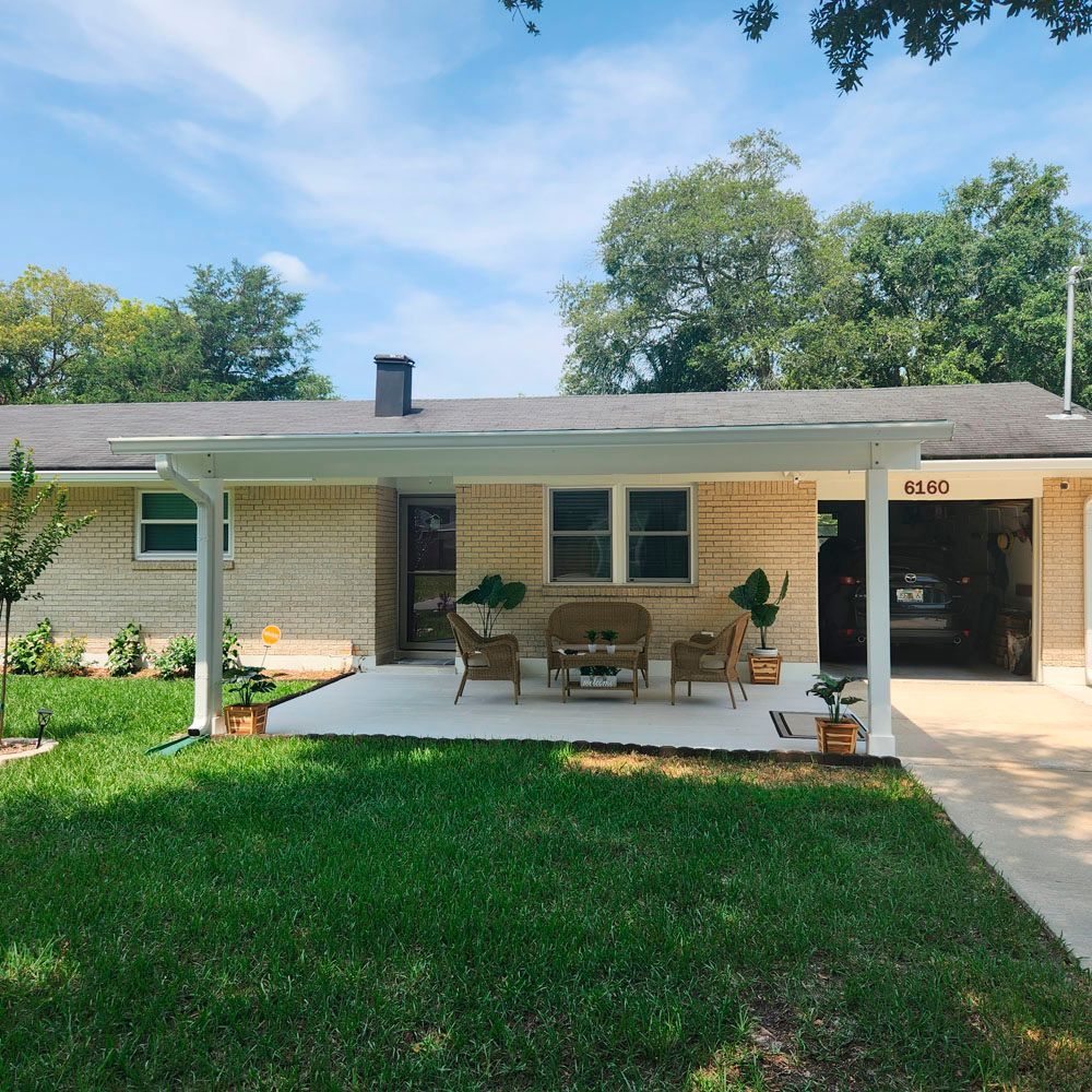 Ranch-style house with patio, green lawn, and open garage. Neutral brick exterior, white patio furniture, sunny day.