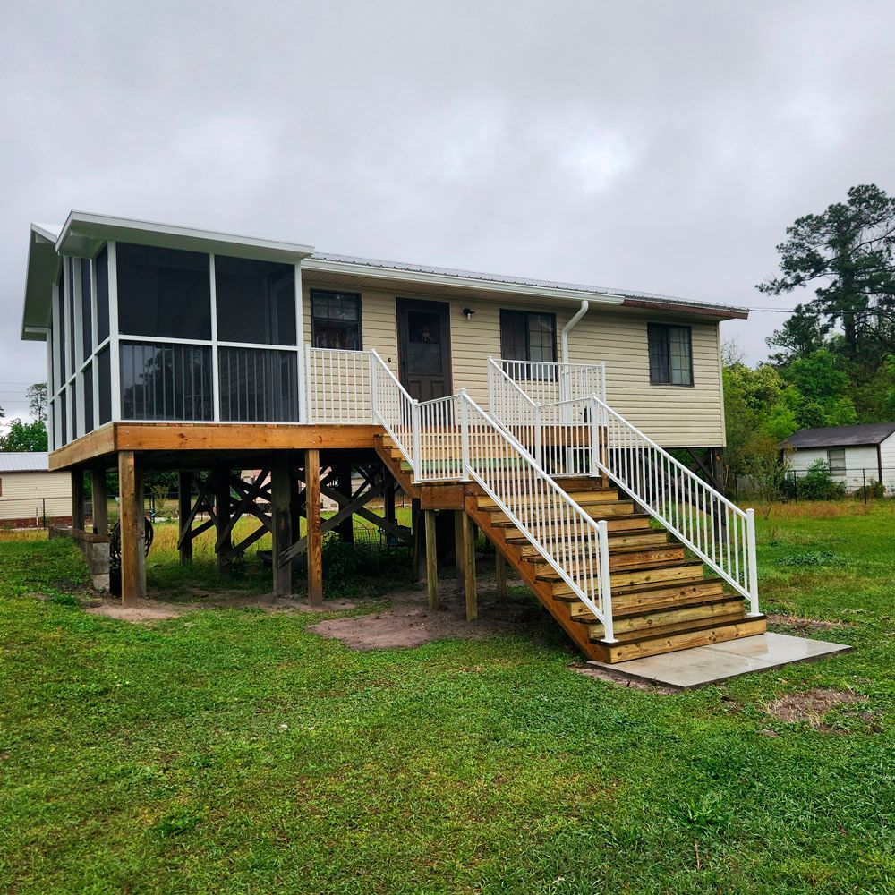 Elevated house with porch and stairs, beige siding, white railings, set on green grass under cloudy sky.