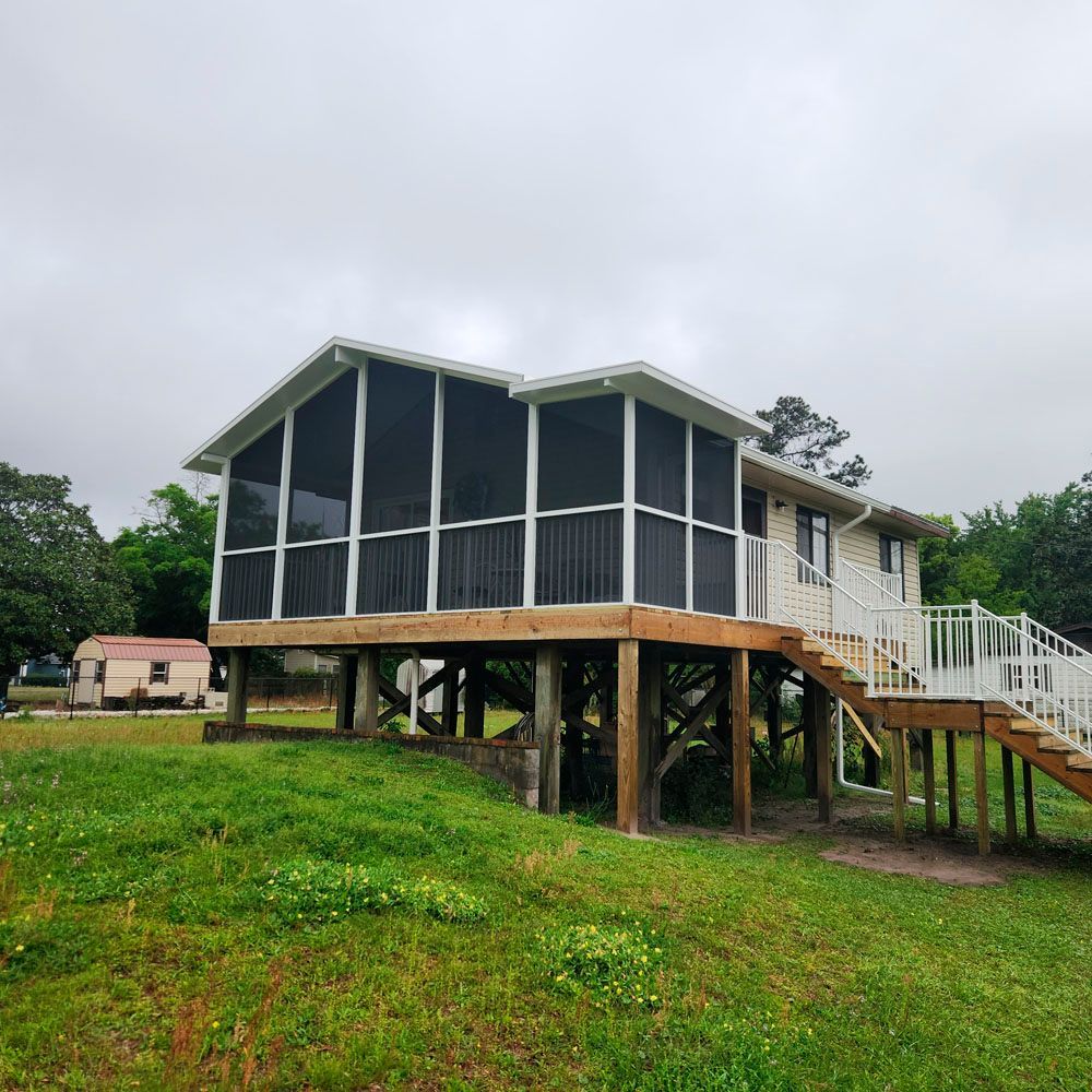 A light-colored house with a screened porch elevated on wooden supports. White railings and a staircase are visible.