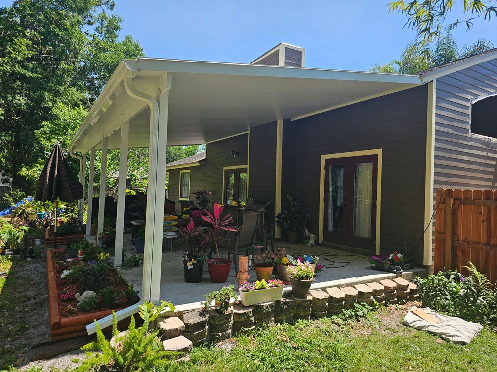 Patio with a white covered roof, next to a brown house with a garden and plants.