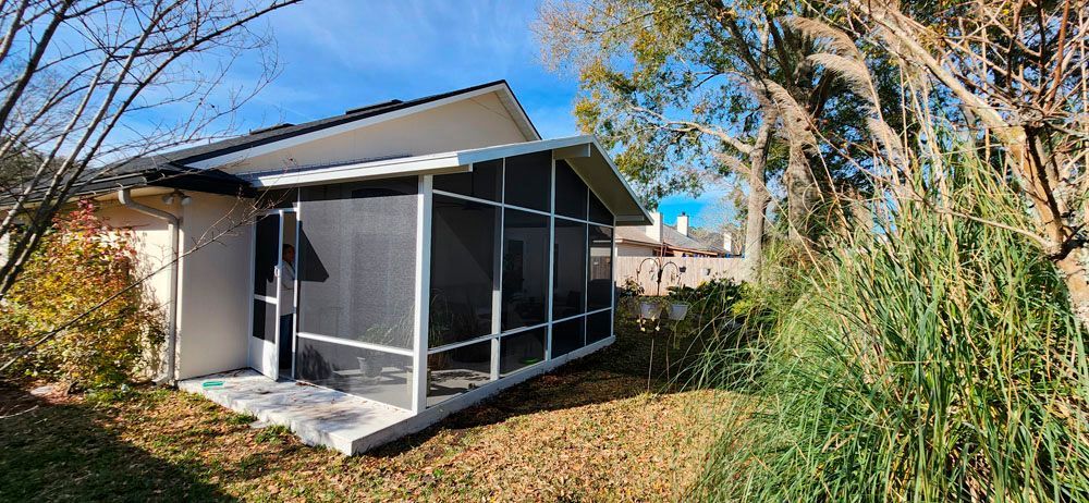 Screened porch attached to a white house with a blue sky background and tall grass in front.