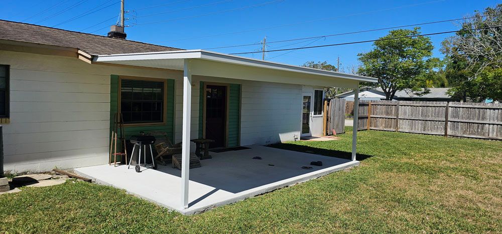 A concrete patio with a white awning extends from a house, with green grass and a wooden fence in the backyard.