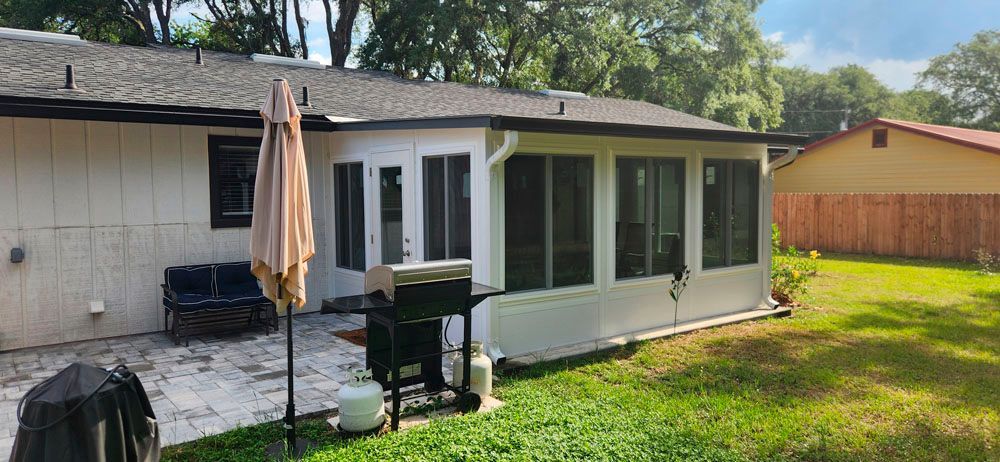 Backyard patio with a screened-in porch and grill. Green grass and trees surround.