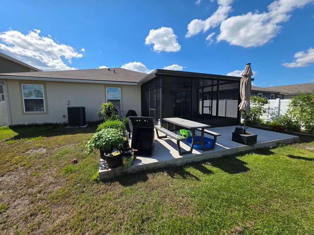 Backyard patio with screened enclosure, picnic table, grill, and potted plants on a sunny day.