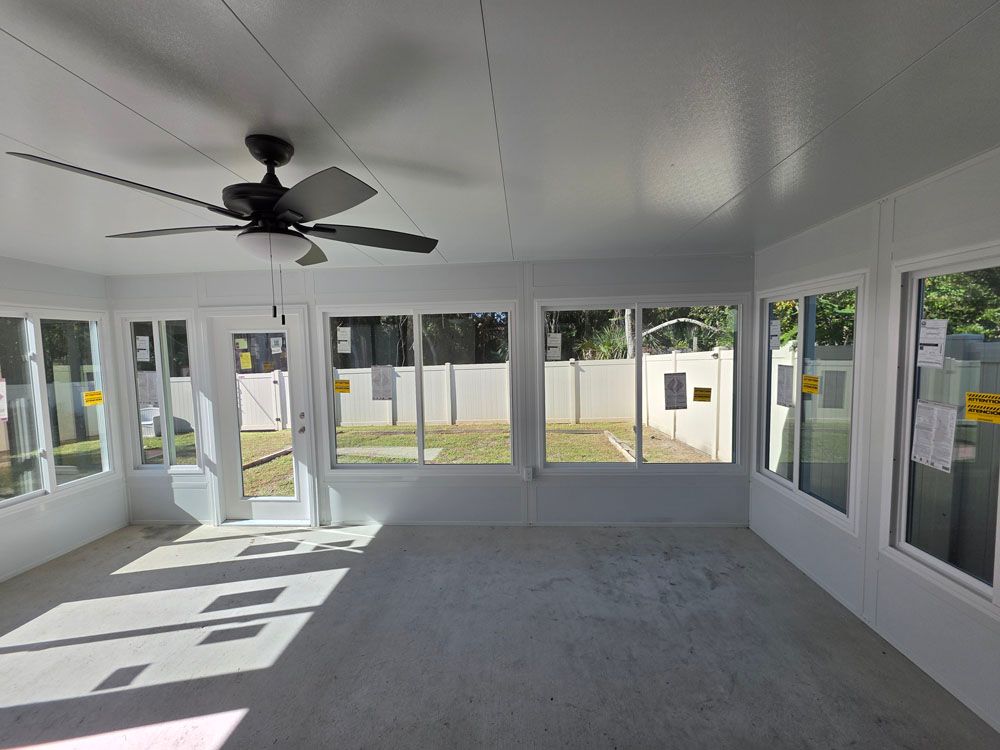 Interior of a sunroom with white walls and windows, a ceiling fan, and concrete floor, overlooking a yard.