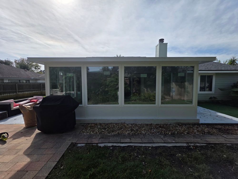 White sunroom with large windows and a chimney, attached to a light green house. A grill and patio furniture are nearby.
