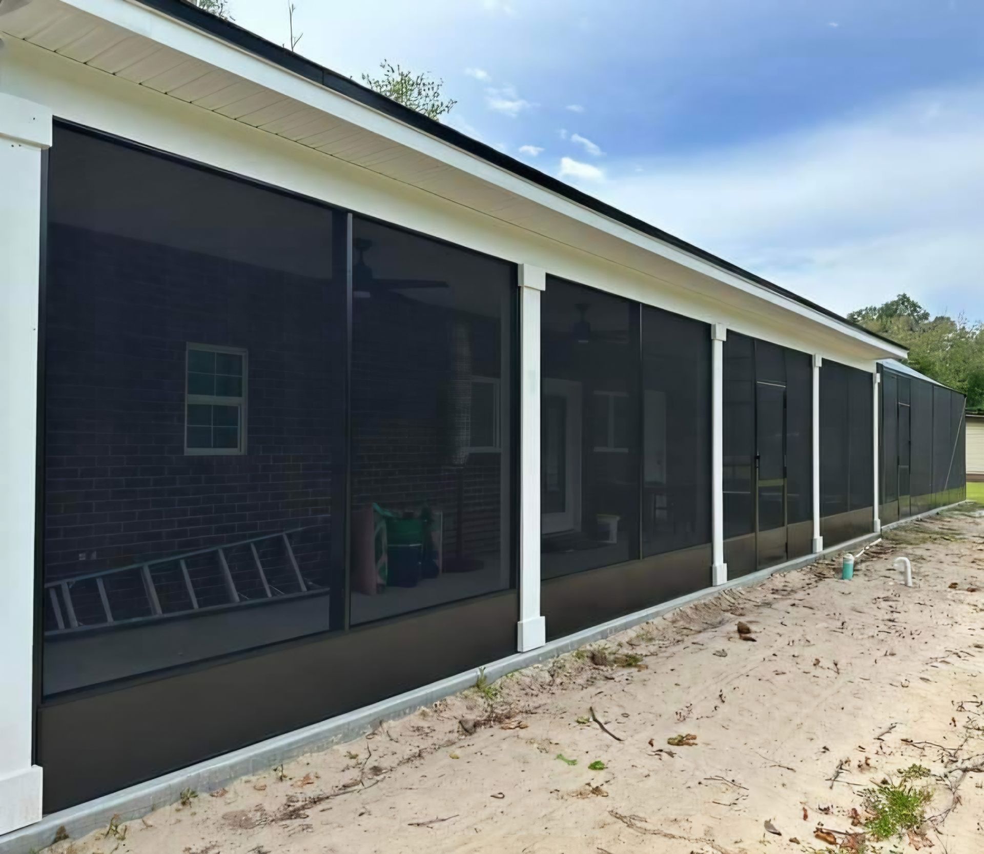 A long screened-in porch with white columns and dark frames exterior to a brick home over a sandy landscape.